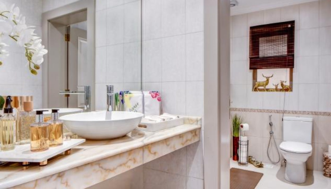 Modern bathroom with a marble countertop sink, various toiletries, a wall mirror, and a separate area with a toilet and decorative items. Bright lighting and neutral tiles throughout.