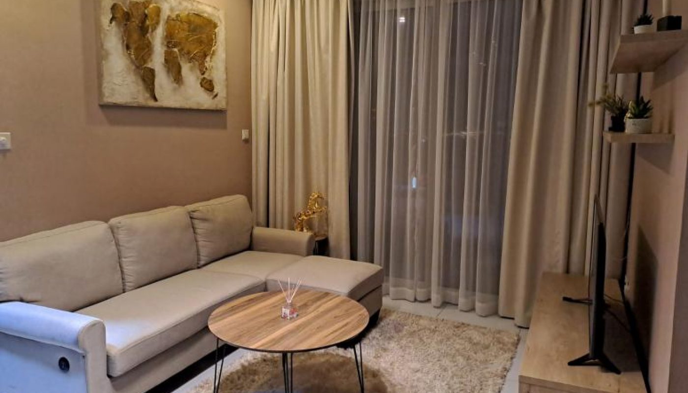 Modern living room with a beige sofa, round wooden coffee table, TV on a wooden stand, wall shelves, world map art, and beige curtains covering a window.
