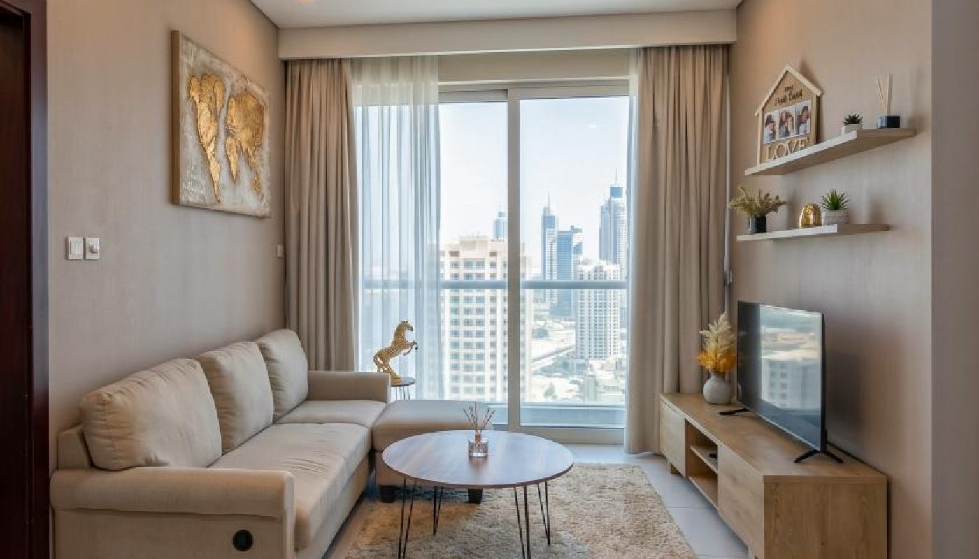 Modern living room with beige sofa, coffee table, rug, TV stand, wall art, shelves, and large window overlooking city buildings. Ceiling light fixture and tile flooring are visible.