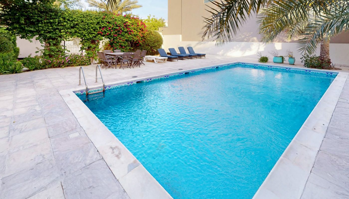 Rectangular outdoor swimming pool with clear blue water, surrounded by marble tiles, palm trees, lounge chairs, and a table with chairs in a private garden setting.