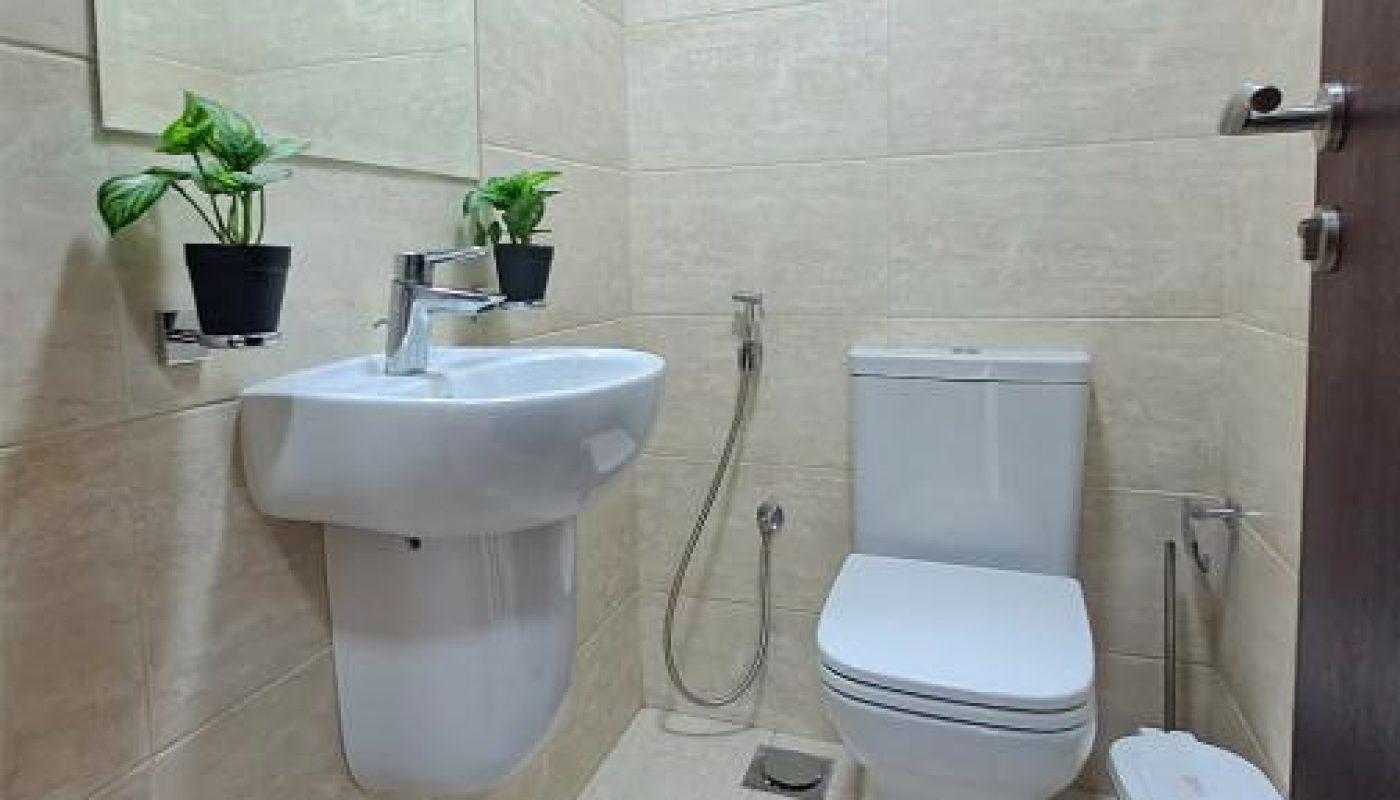 A small bathroom with a wall-mounted sink, a toilet, two potted plants, a mirror, and a handheld bidet sprayer on beige tiled walls and floor.