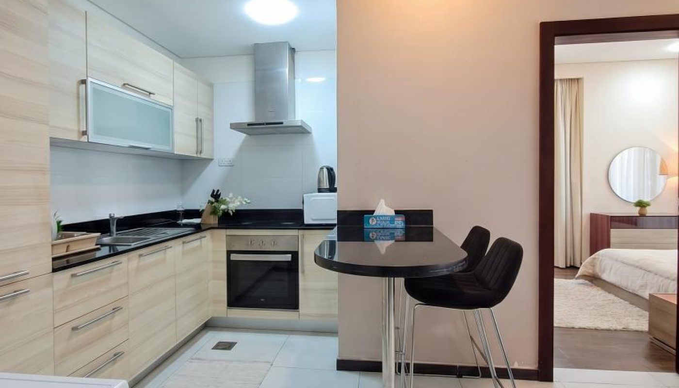 Modern apartment kitchen with light wood cabinets, black countertop, small dining area with two black chairs, and a view into a bedroom with a bed and round mirror.