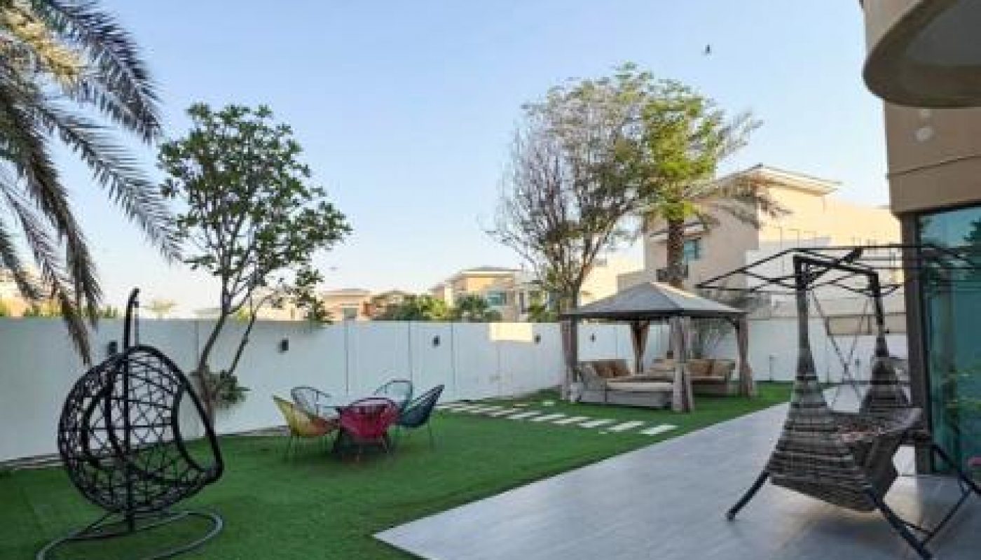 A backyard with artificial grass, lounge chairs, a hanging egg chair, patio swing, gazebo, and trees, bordered by a white wall and nearby houses under a clear sky.