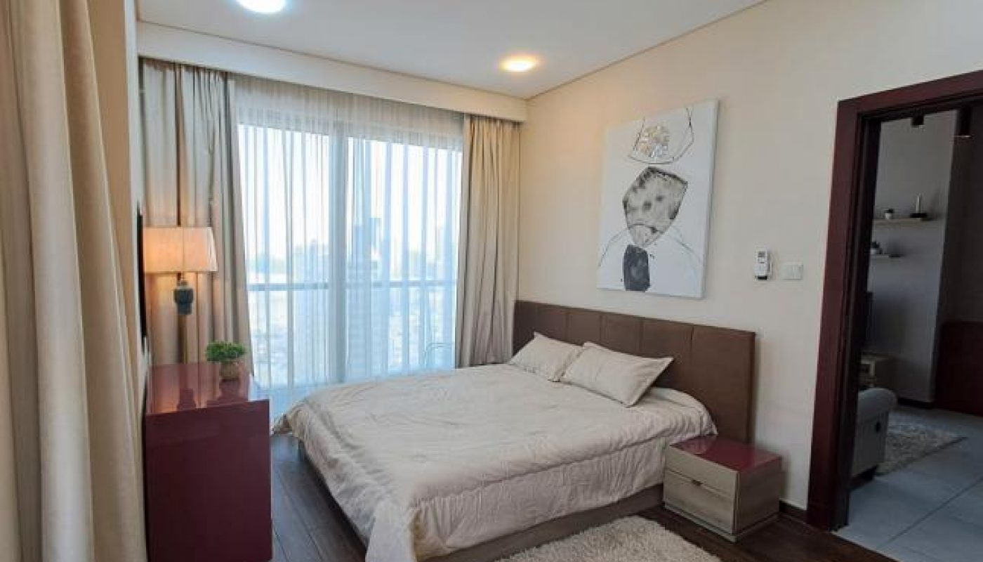 A modern bedroom with a double bed, beige bedding, a nightstand, a lamp, a large rug, floor-to-ceiling curtains, and a portrait painting above the bed.