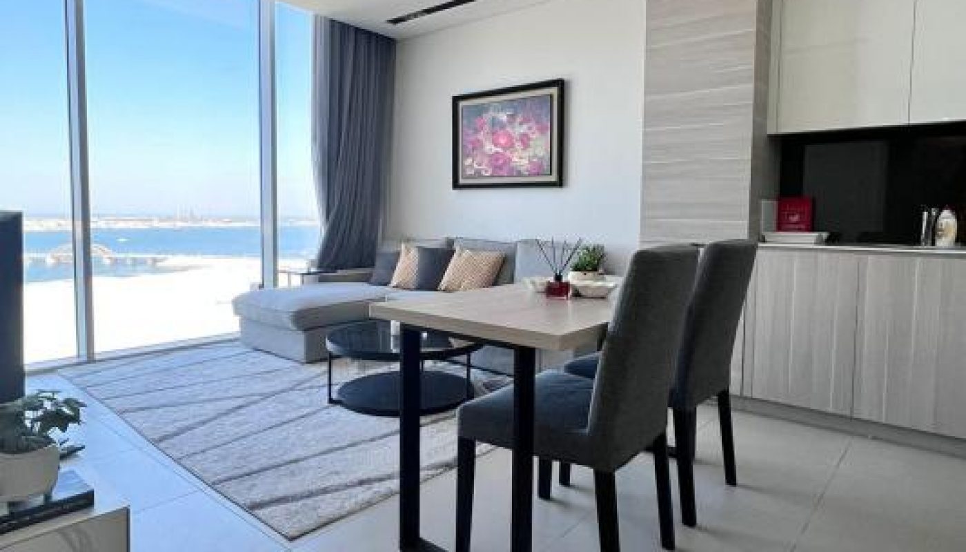 Modern apartment with dining table, chairs, and sofa near large floor-to-ceiling windows with a city view; kitchen area on the right and a framed painting on the wall.