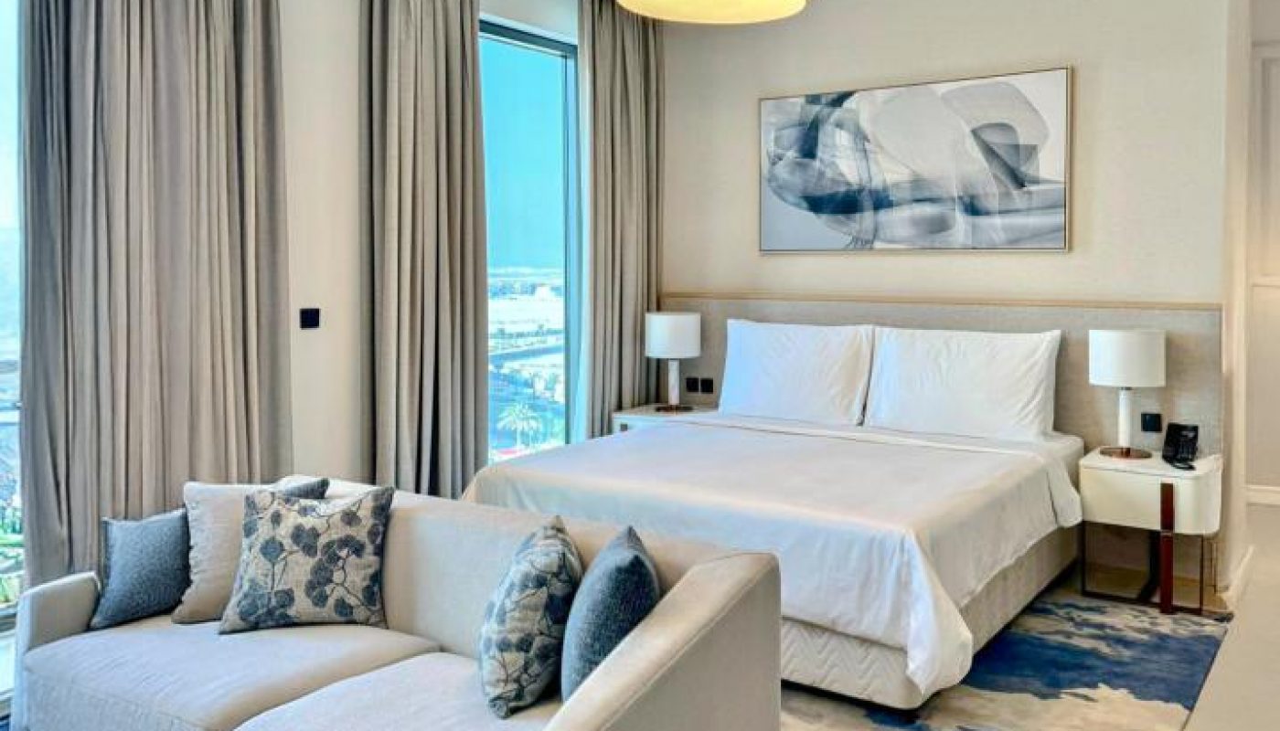 Modern bedroom with large windows, a double bed, a light sofa at the foot of the bed, blue and white rug, and neutral decor.