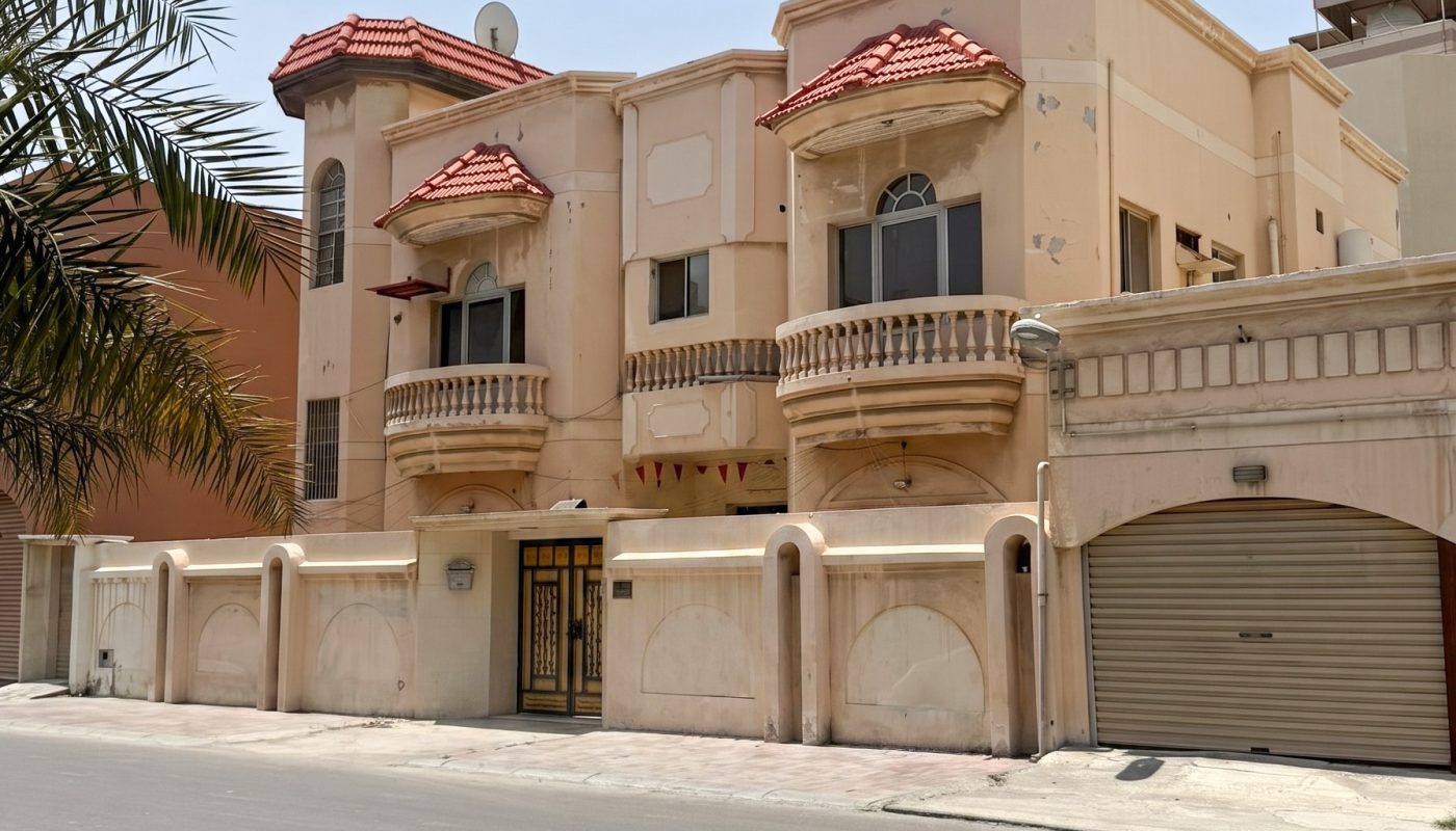 A beige two-story house with red-tiled roof accents, arched windows, and balconies, surrounded by a high wall and closed gates, on a quiet street under a clear sky.