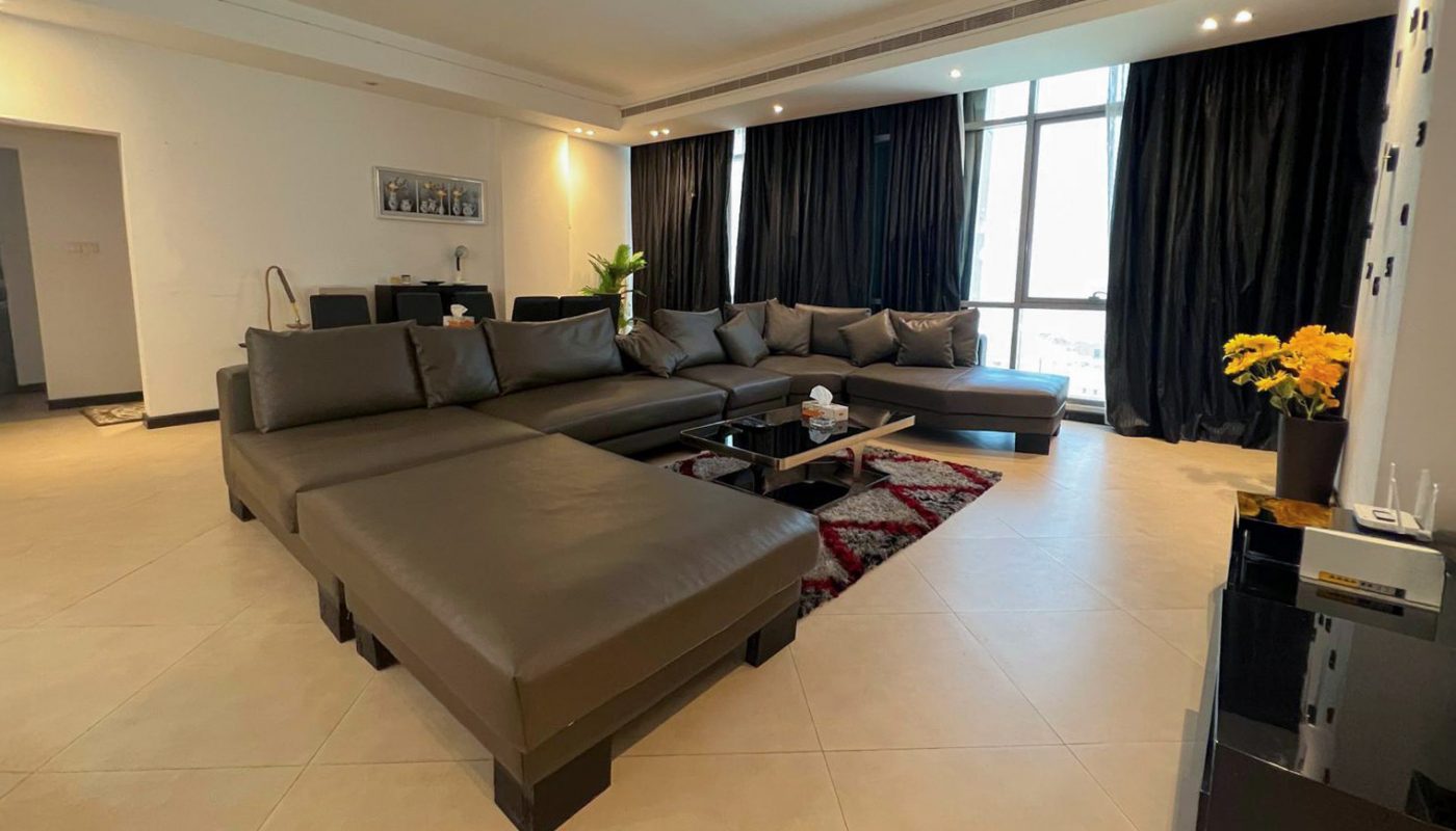 Modern living room with a large sectional sofa, black coffee table, red rug, floor-to-ceiling windows with black curtains, and a corner table with yellow flowers.