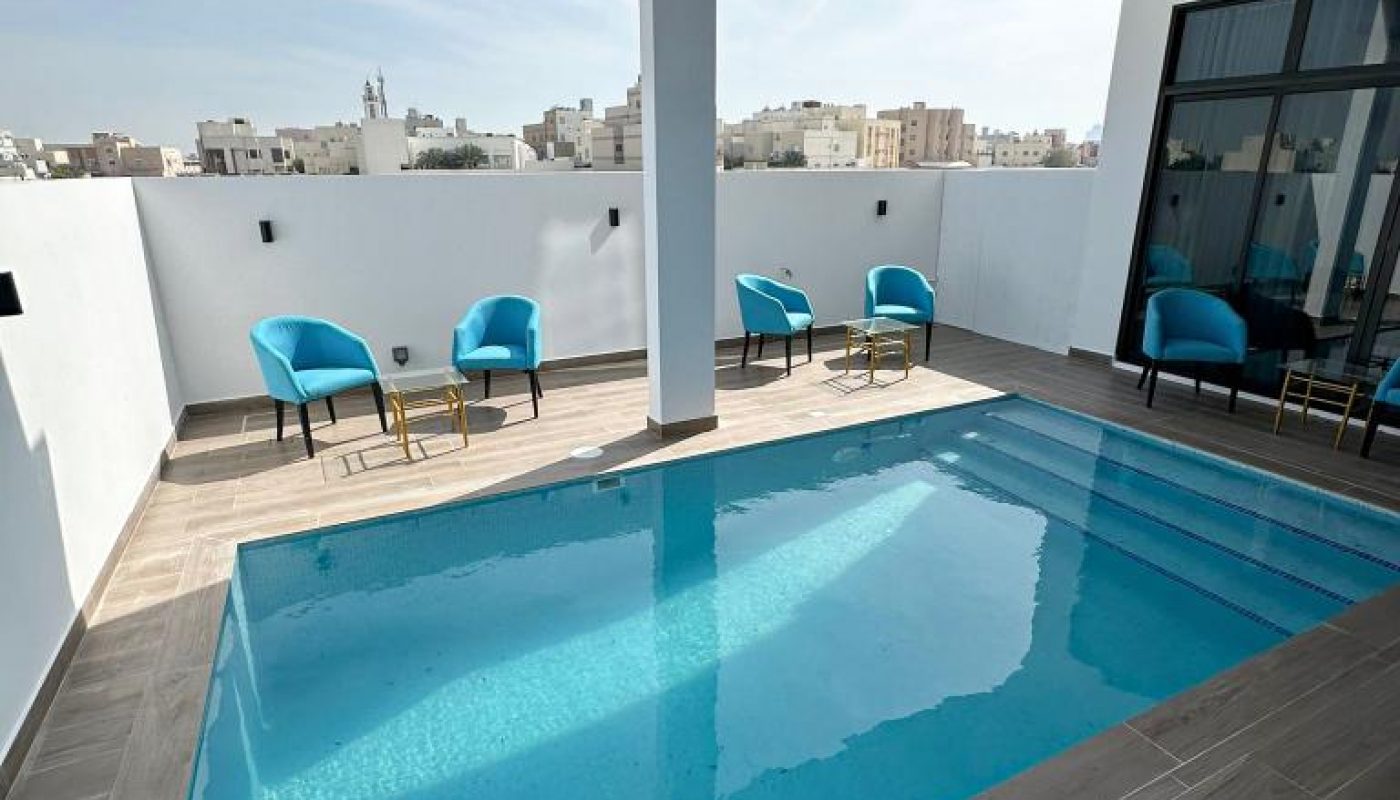 A small rectangular outdoor pool with turquoise water is surrounded by gray tile flooring and blue chairs, set against white walls under a partly covered patio.
