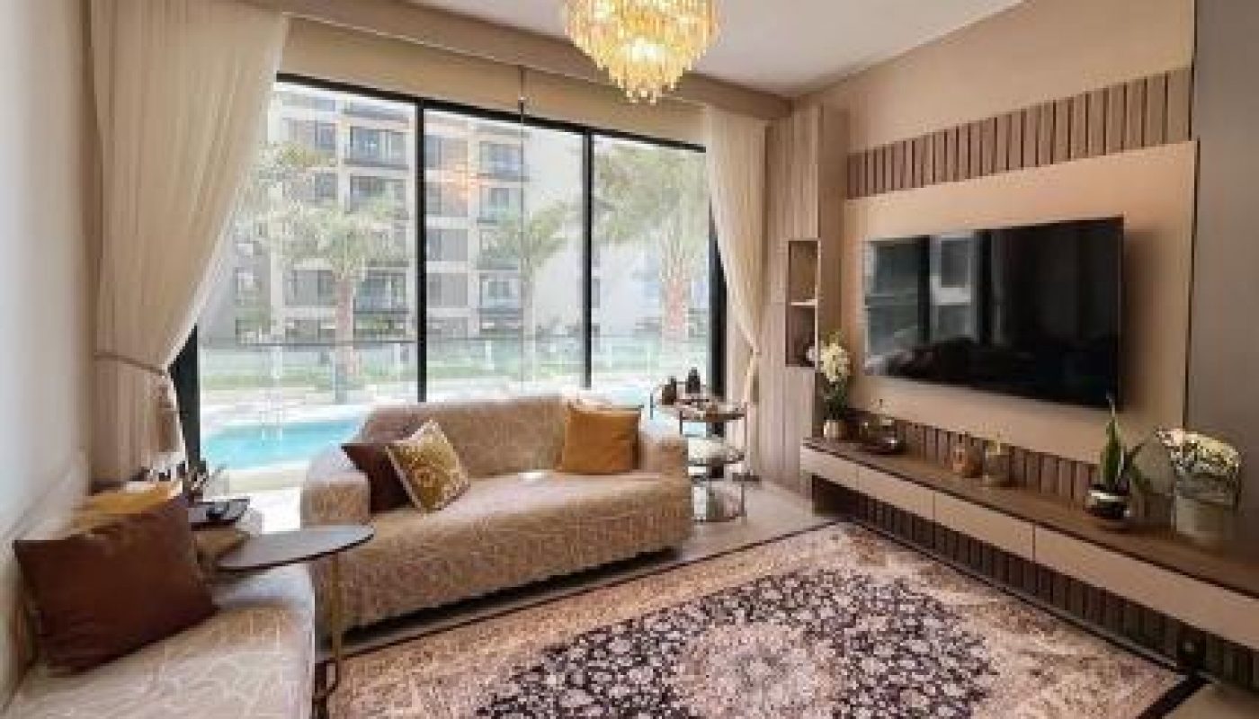 Modern living room with a patterned rug, sofa, wall-mounted TV, large window with pool view, and a chandelier overhead.