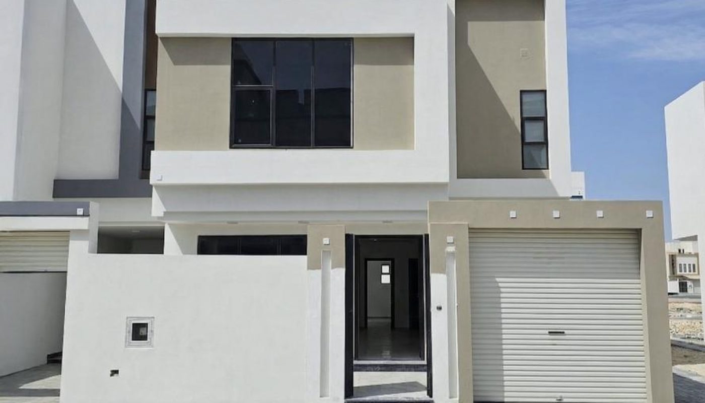 A modern two-story house with a flat roof, large windows, a garage with a closed white door, and a white and beige exterior under a blue sky with wispy clouds.
