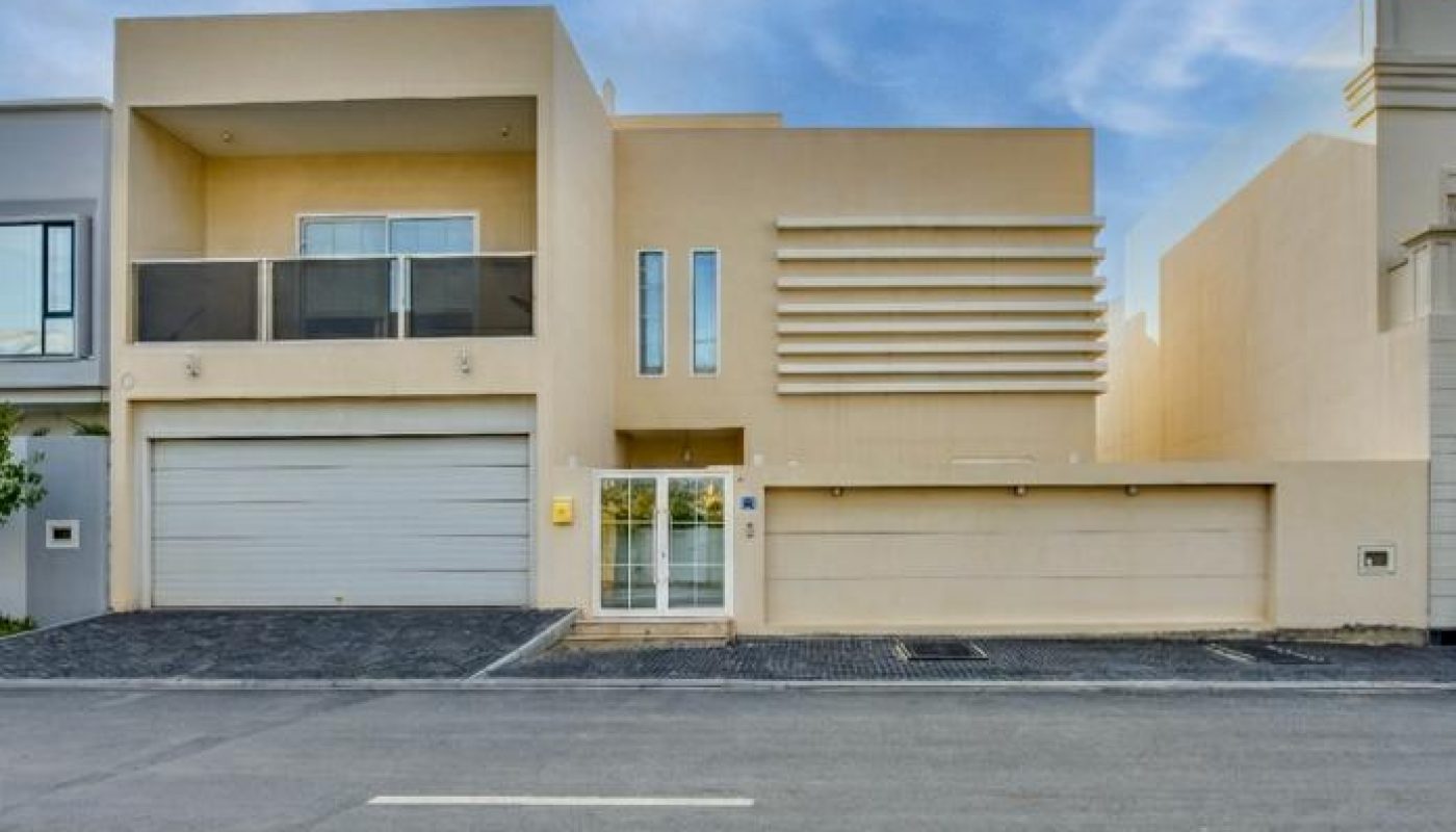 Modern beige two-story house with large garage doors, balcony, and flat roof, set on a paved street under a partly cloudy sky.
