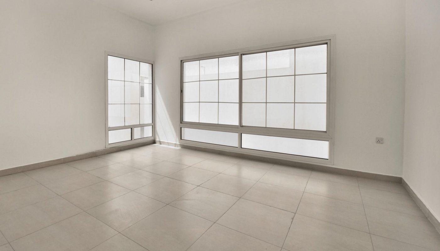 Empty, unfurnished room with large white-tiled floor, plain white walls, and two sets of wide windows letting in natural light.