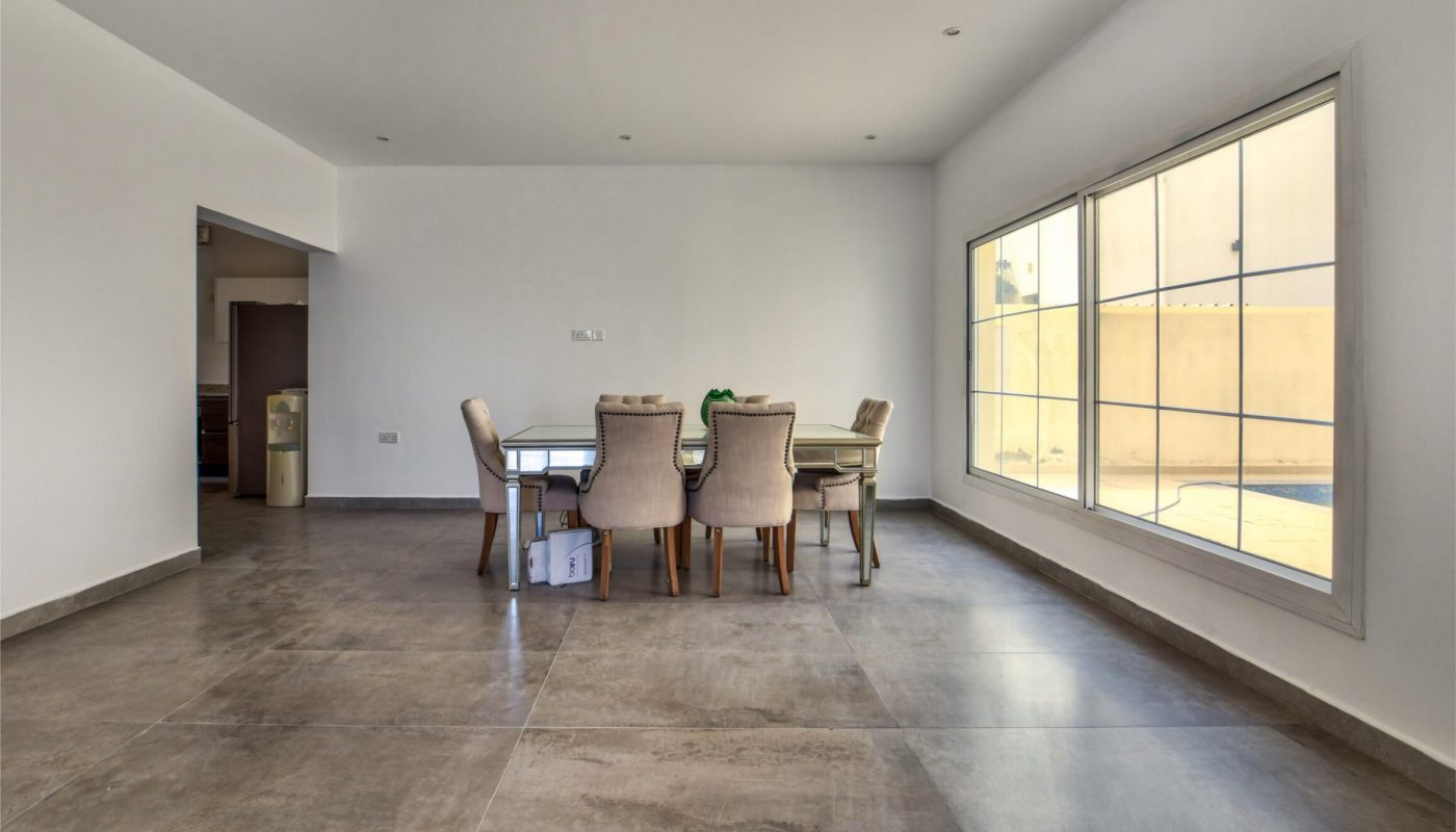 A spacious dining room with a rectangular table and six chairs, large tiled floor, white walls, and a wide window letting in natural light.