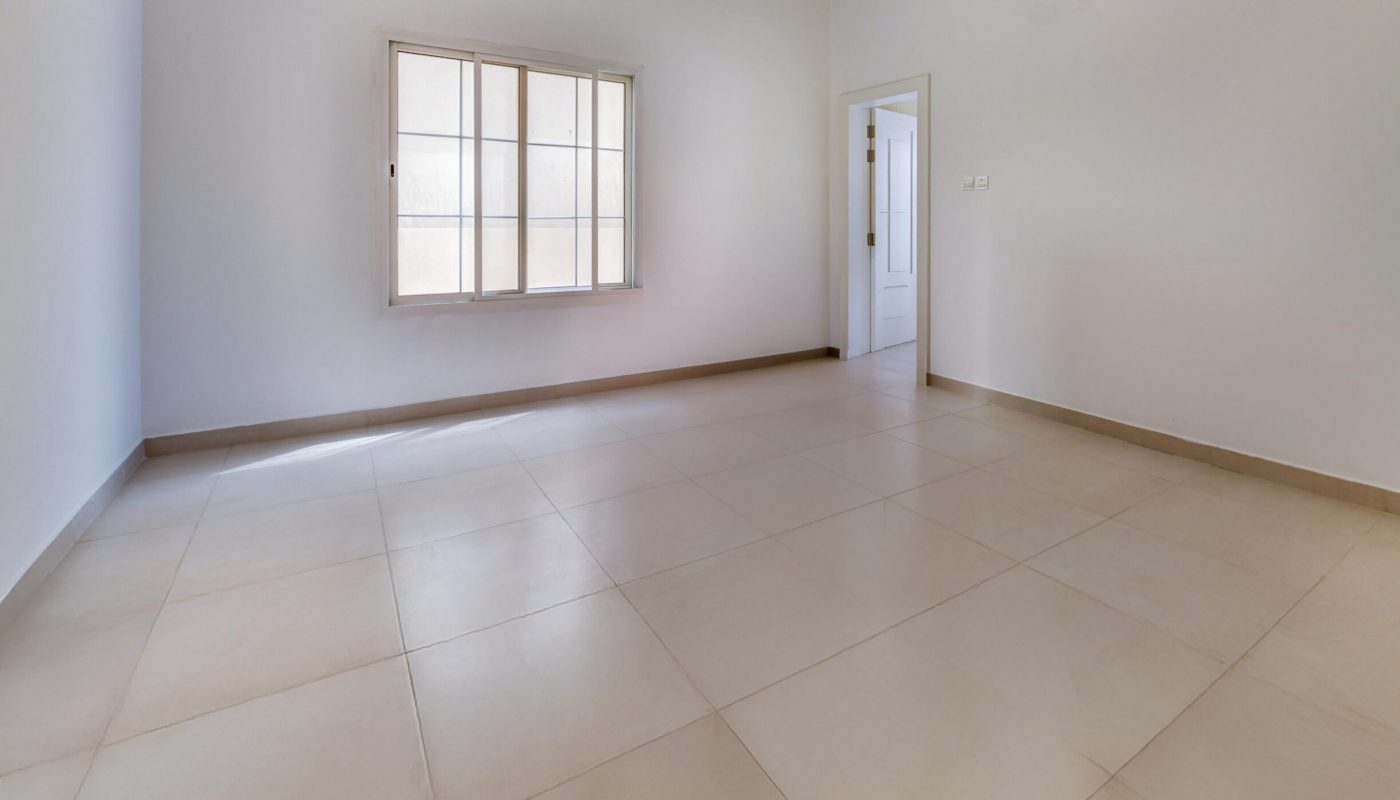 Empty room with large beige floor tiles, plain white walls, a window with white framing, and an open doorway leading to another area.