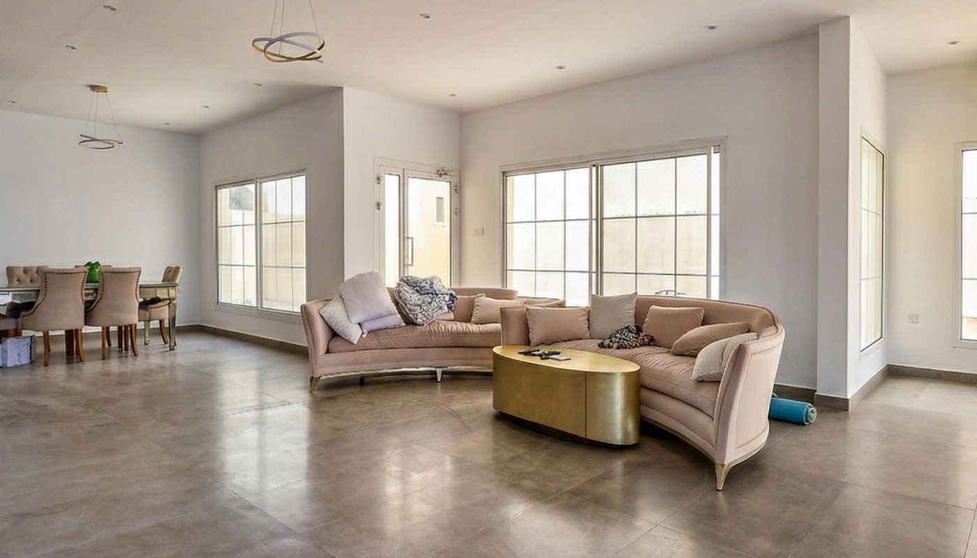 Spacious, unfurnished living room with large windows, beige sofas, a gold coffee table, and an adjacent dining area with a table and chairs.