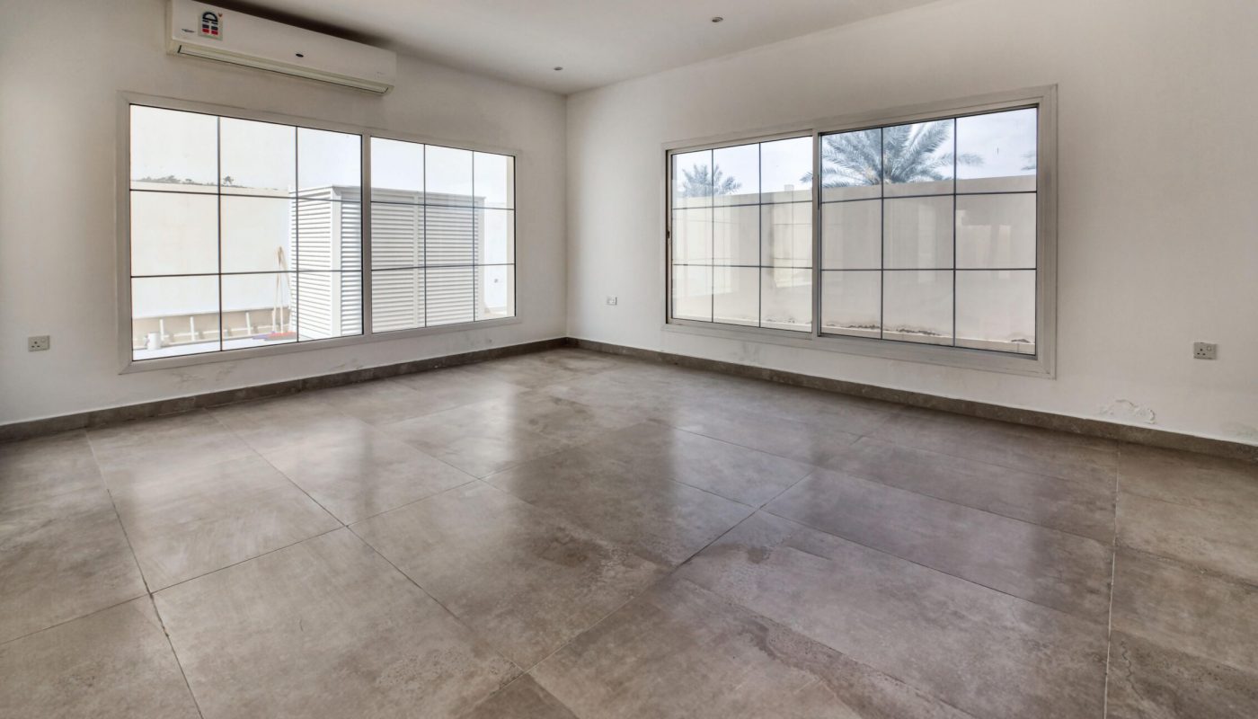 An empty room with large tiled floors, white walls, two large windows, and a wall-mounted air conditioning unit.