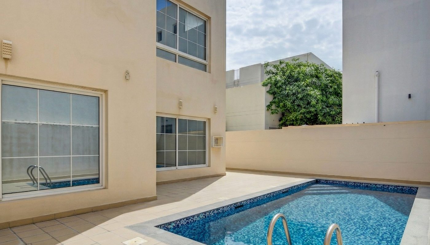 Rectangular swimming pool surrounded by beige tiled patio, adjacent to a modern building with large windows and a privacy wall under a partly cloudy sky.