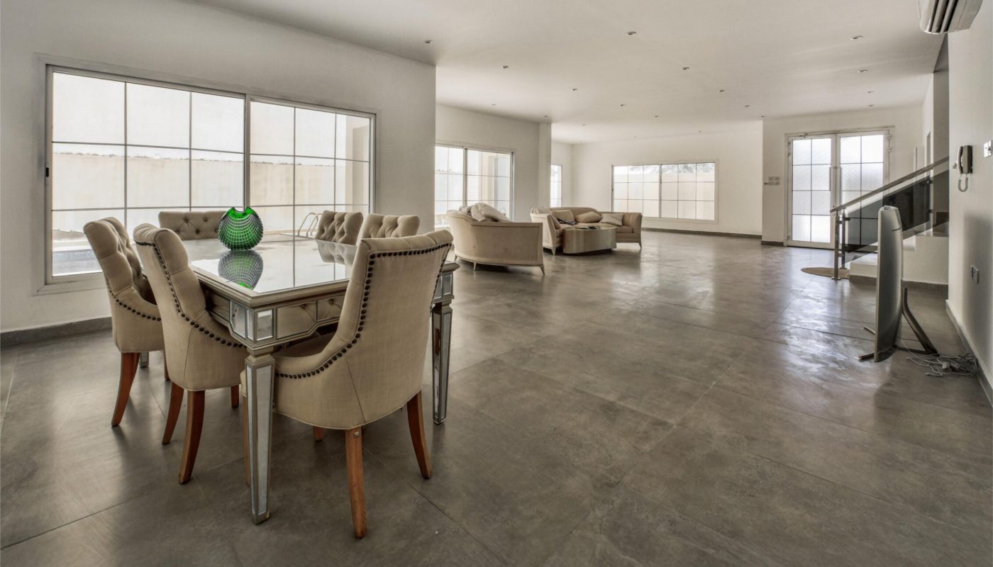 Spacious, modern living and dining area with large windows, light-colored walls, beige furniture, and a concrete floor.