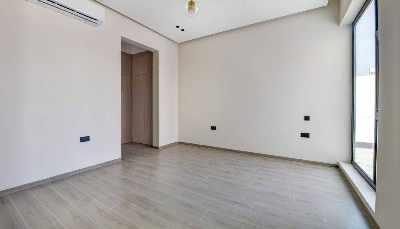 Empty modern room with light wood flooring, beige walls, a ceiling light fixture, built-in closet, wall-mounted air conditioner, and large window letting in natural light.