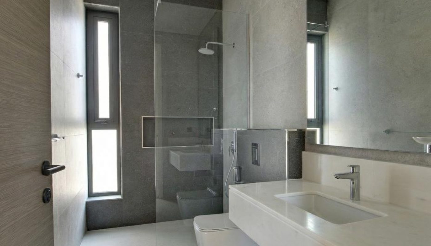 Modern bathroom with gray walls, large mirror, wall-mounted sink and toilet, and a glass-enclosed shower next to a tall window.