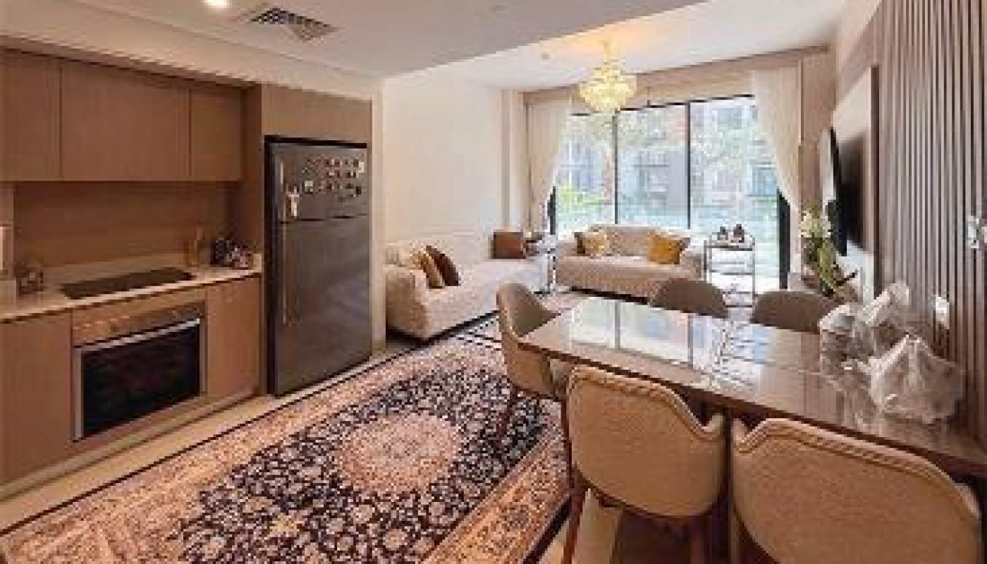 Modern open-plan kitchen and living area with a dining table, beige sofa, large patterned rug, and floor-to-ceiling windows with curtains.