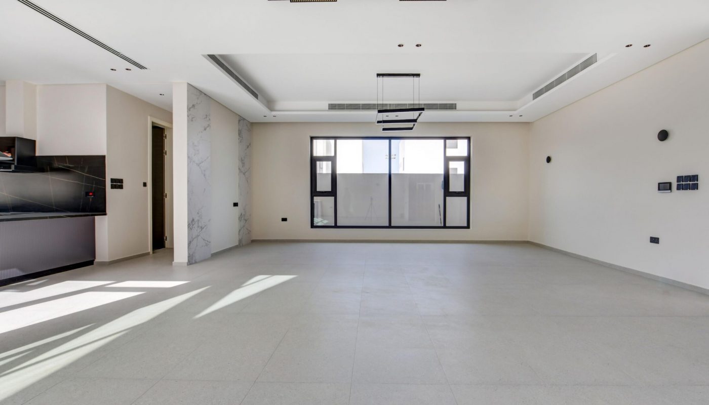 Spacious empty modern living room with large windows, minimalistic lighting fixtures, tiled flooring, and an open view of the kitchen area.