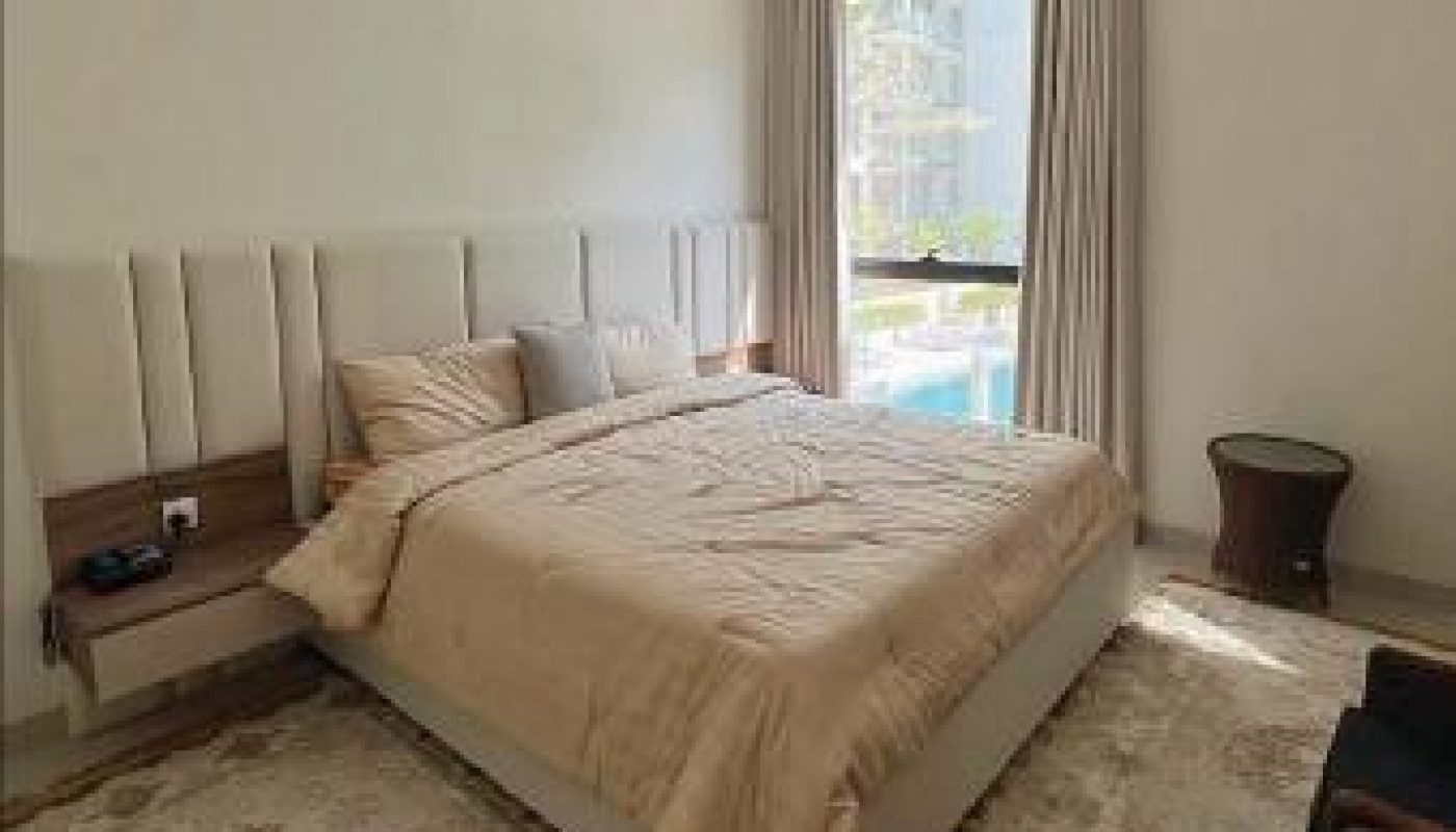A neatly made bed with beige bedding is centered in a bright bedroom with two rugs, a chandelier, a window with curtains, and a small table.