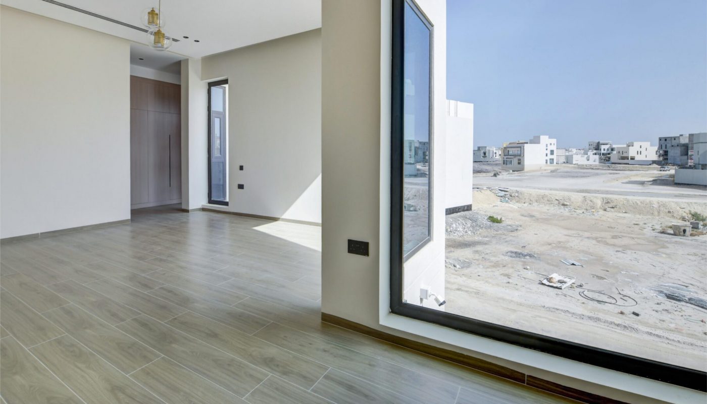 Minimalist empty room with large floor-to-ceiling window, light wood tile flooring, and views of a modern residential development under a clear sky.