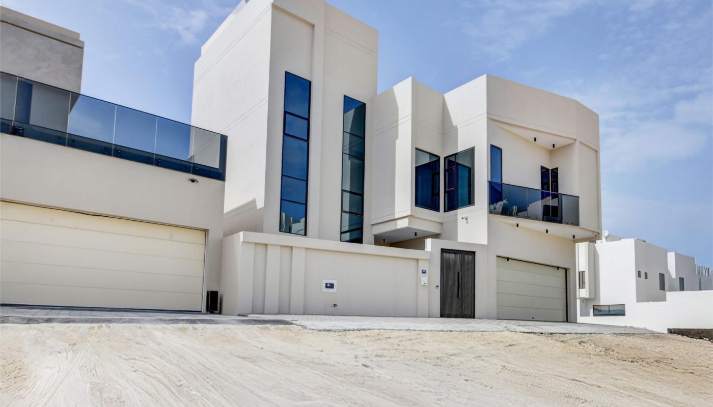 Modern white two-story house with large windows, glass balconies, and multiple garages, situated on a sandy lot under a partly cloudy sky.
