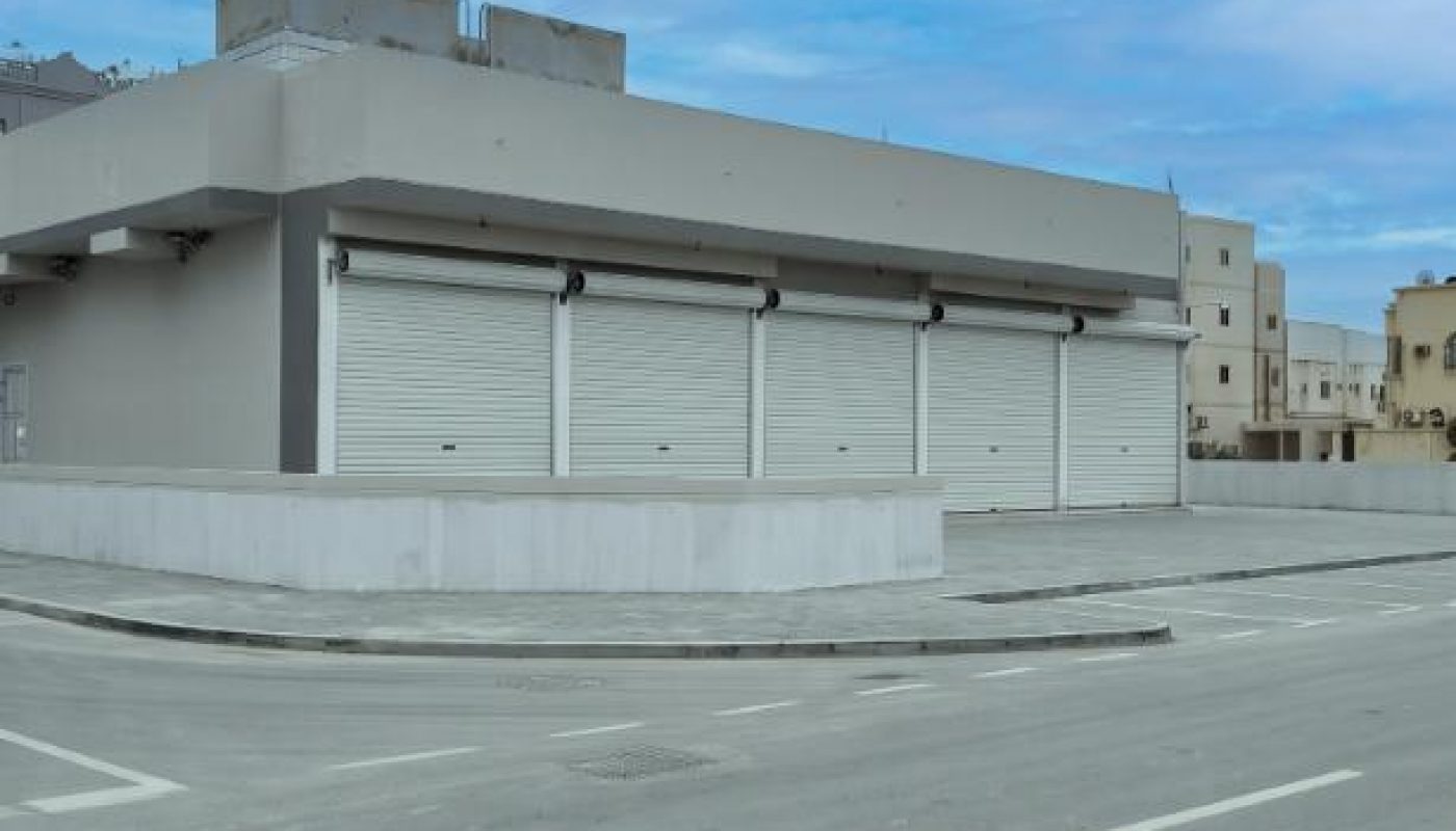 A gray commercial building with three closed metal shutters faces an empty street under a blue sky with scattered clouds.