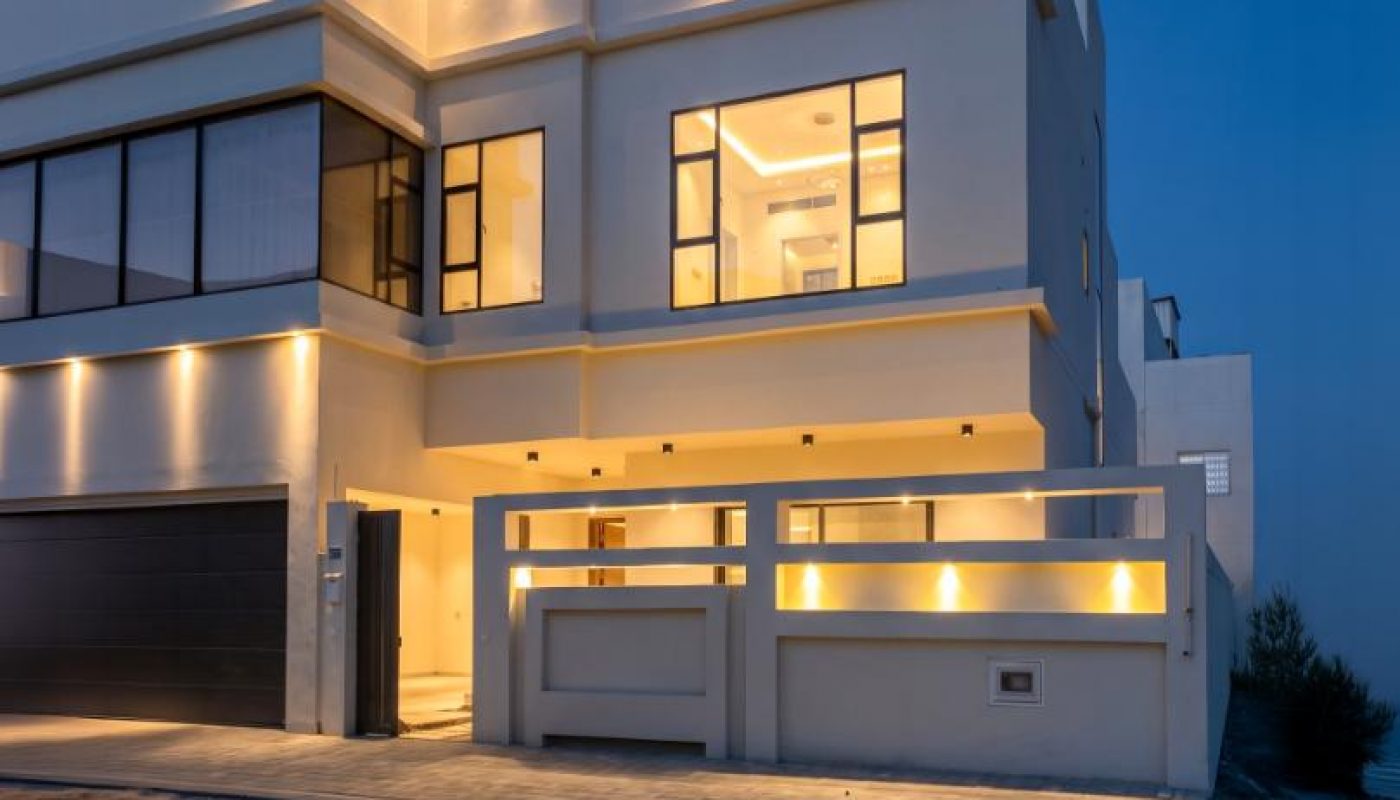 A modern two-story house with large windows and exterior lights on, viewed at dusk with a sandy driveway in front.