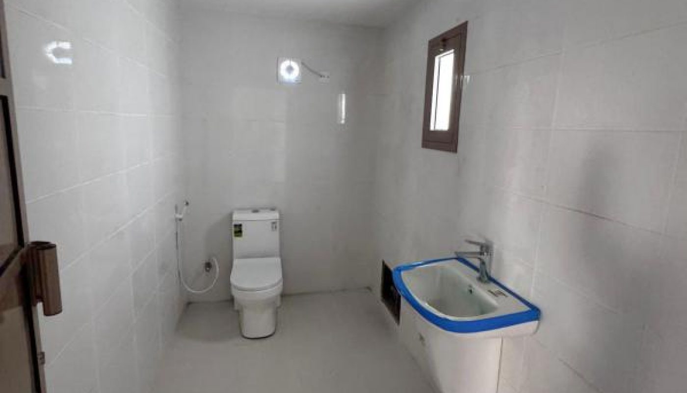A clean, minimal bathroom with white walls and floor, a toilet, a small sink with blue trim, a window, and a handheld bidet spray.