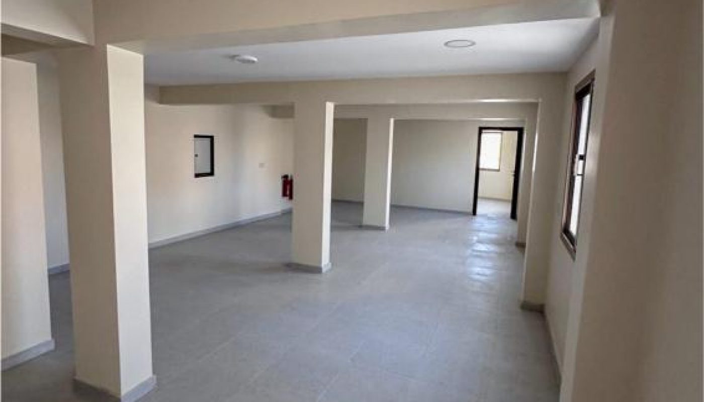 A spacious, empty room with beige walls, tiled floors, multiple support columns, and several windows and doors visible.