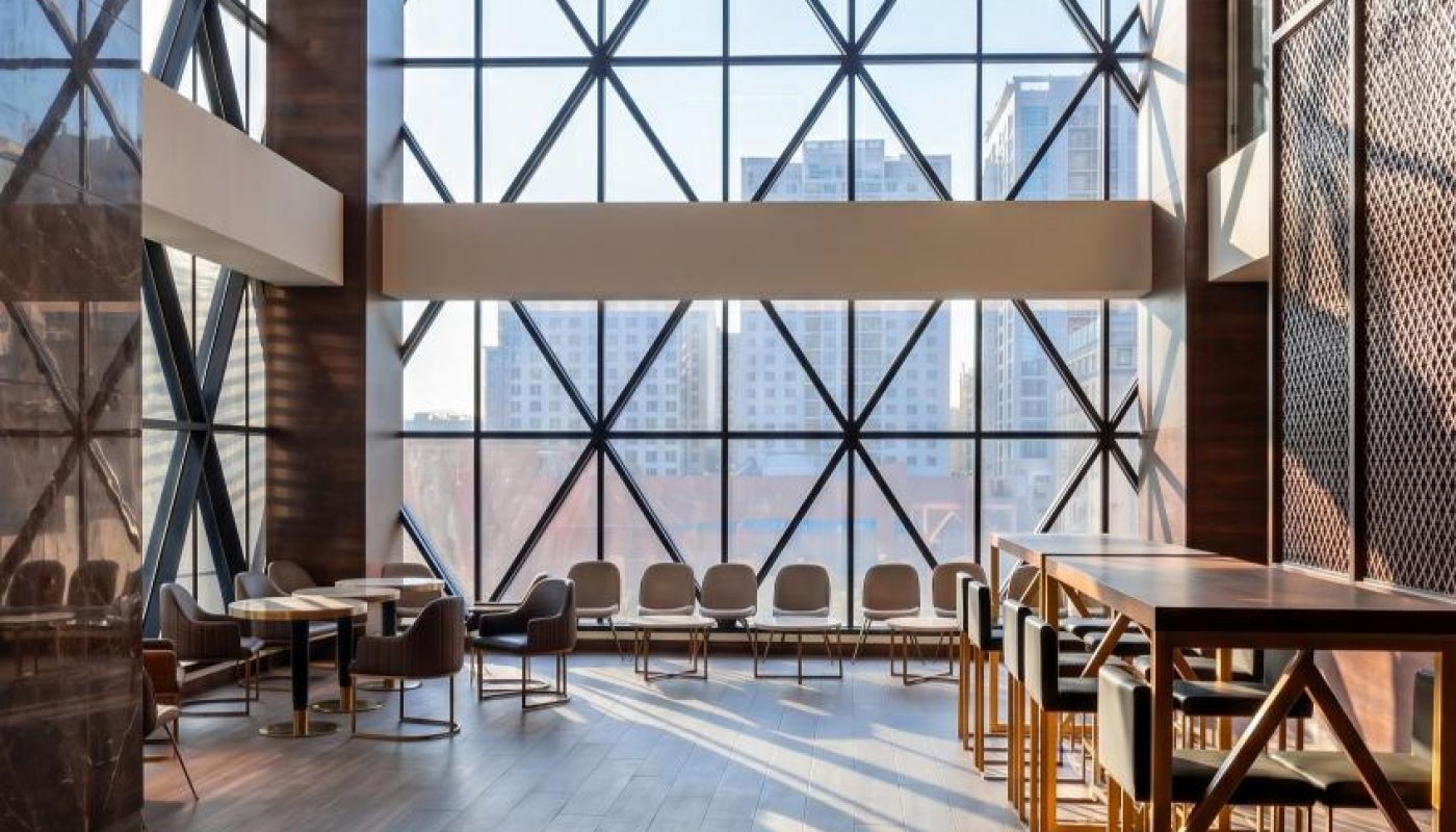Modern waiting area with wooden floors, geometric window panels, and contemporary seating, overlooking a city skyline through large floor-to-ceiling windows.