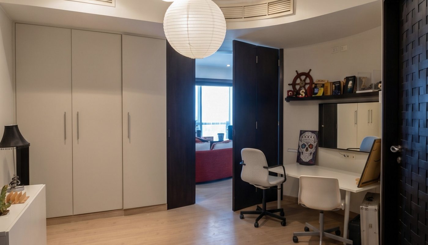 A modern home office with two white chairs at a desk, built-in shelves, closed cabinets, wooden floors, and two round ceiling lights. An open door reveals another room.