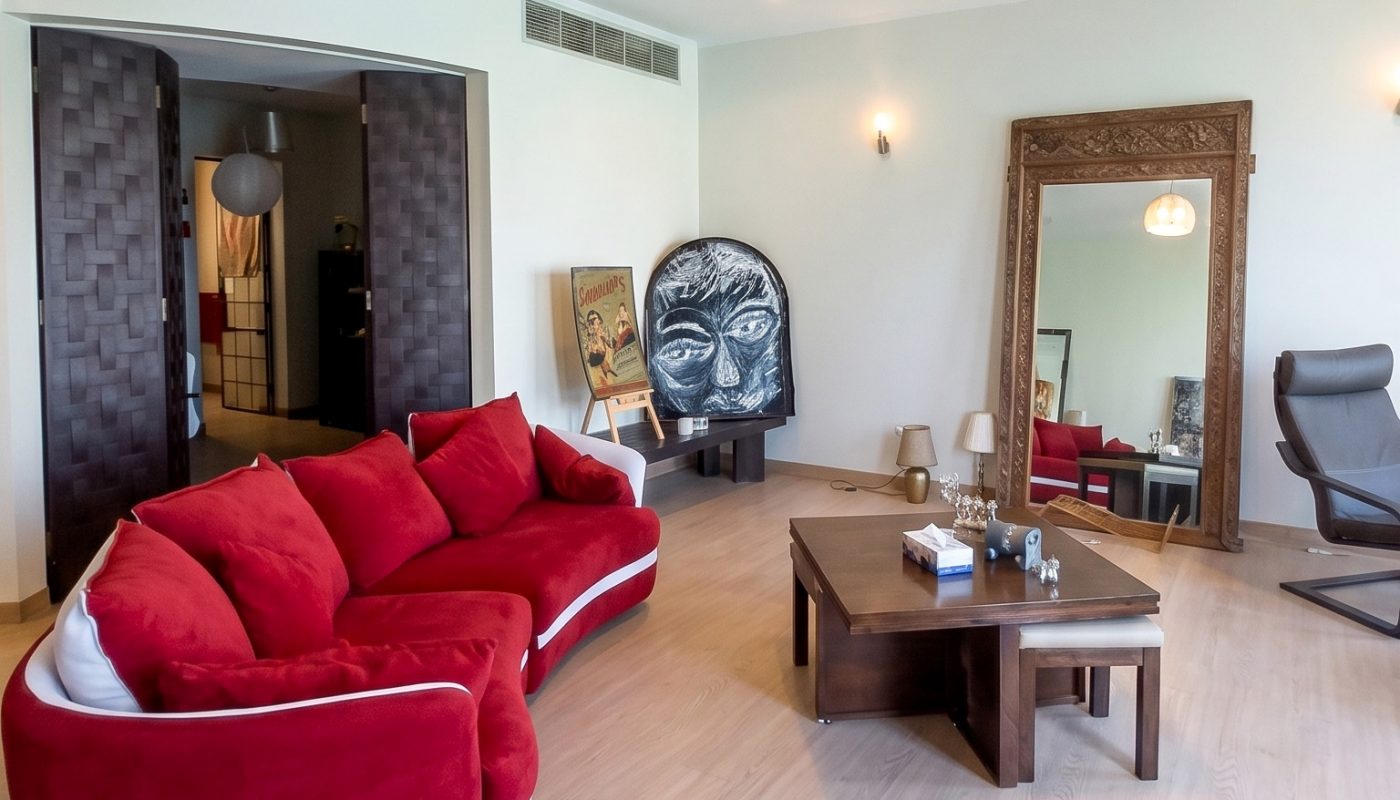A modern living room with a red sofa, a large framed mirror, a wooden coffee table, a gray armchair, and a painting with a face leaning against the wall.