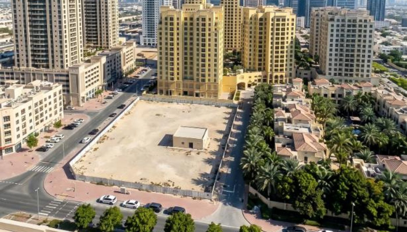 Aerial view of a cityscape showing tall residential buildings, a vacant lot, tree-lined streets, and parked cars under a clear blue sky.