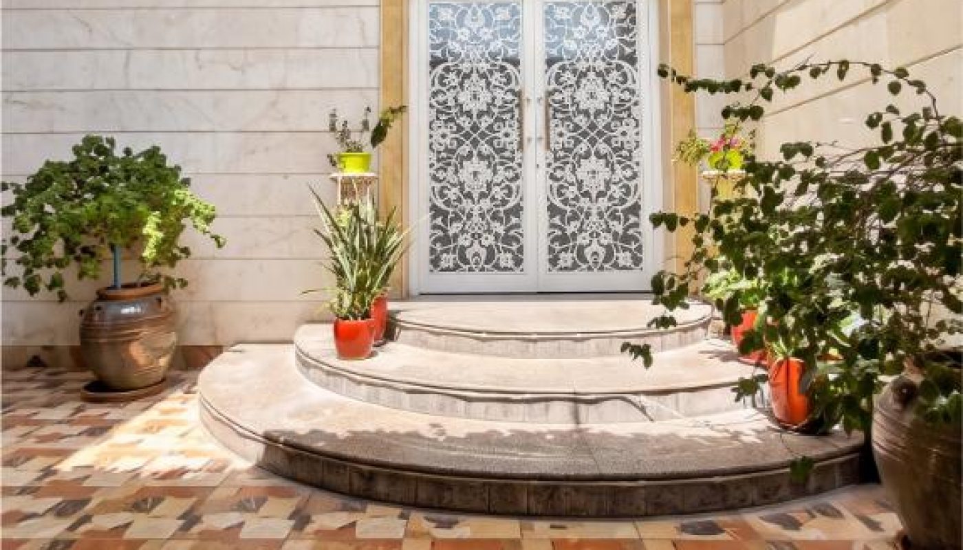 Double doors with ornate glass panels are framed by marble walls and curved steps, surrounded by potted plants on a patterned tile floor under leafy vines.