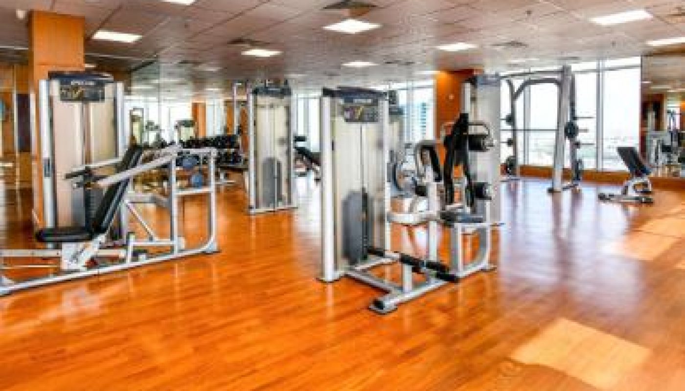 A spacious gym with wooden floors, various weight machines, and large windows letting in natural light.