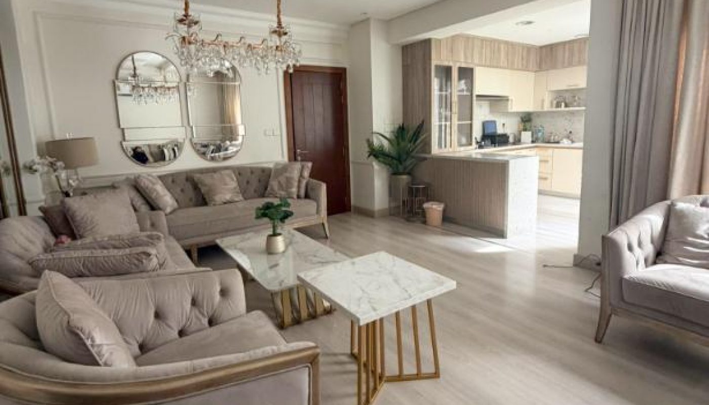 A bright living room with beige armchairs and sofa, a marble coffee table, a chandelier, and an open view into a modern kitchen with light wood cabinets.