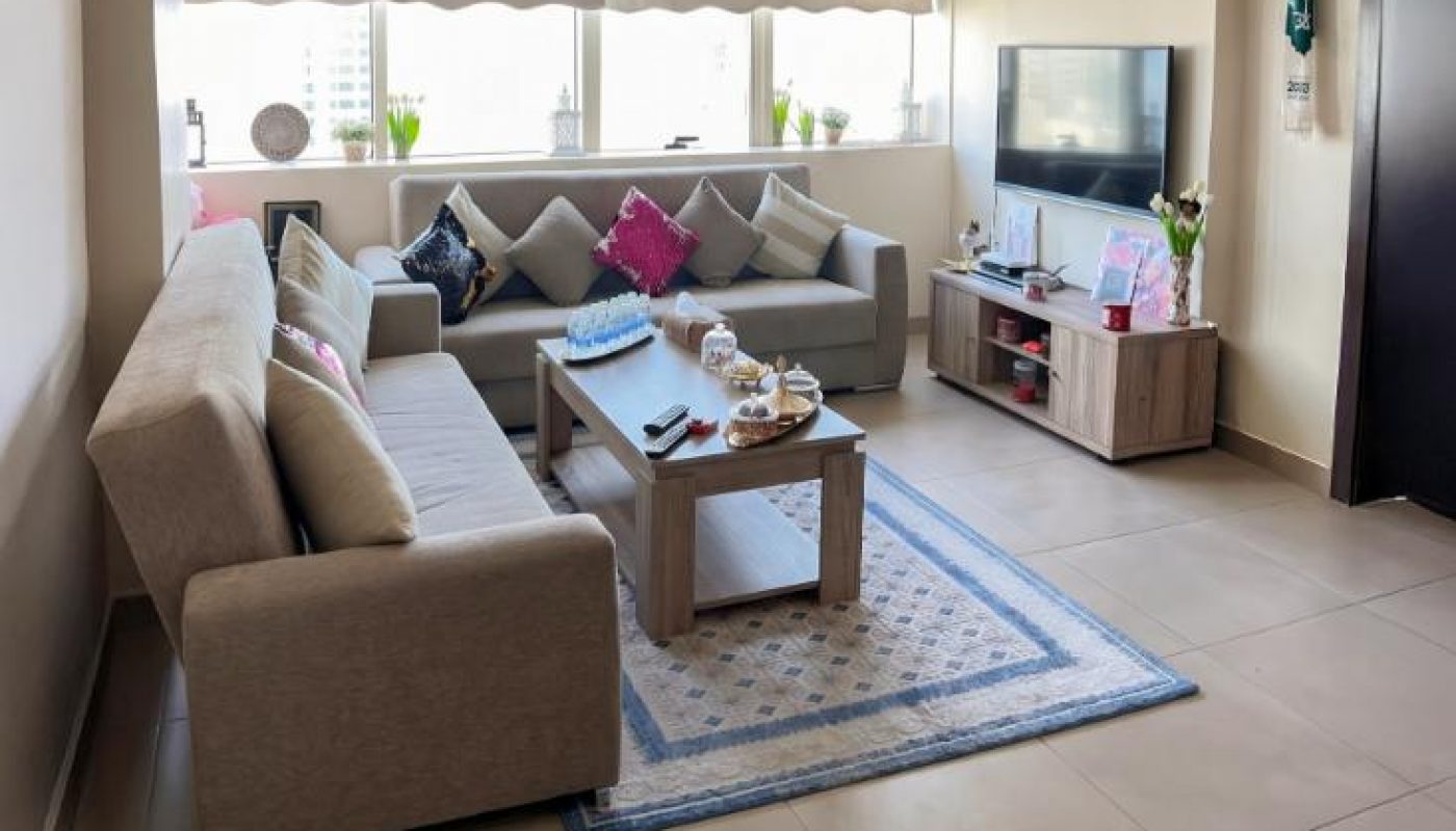 Modern living room with beige sofas, a wooden coffee table, a blue-patterned rug, TV on a stand, and large windows with blinds letting in natural light.
