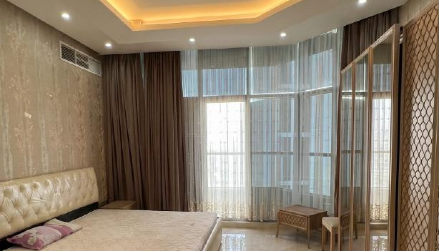 A modern bedroom with a large bed, mirrored wardrobe, two stools, floor-to-ceiling windows, and ceiling recessed lighting.