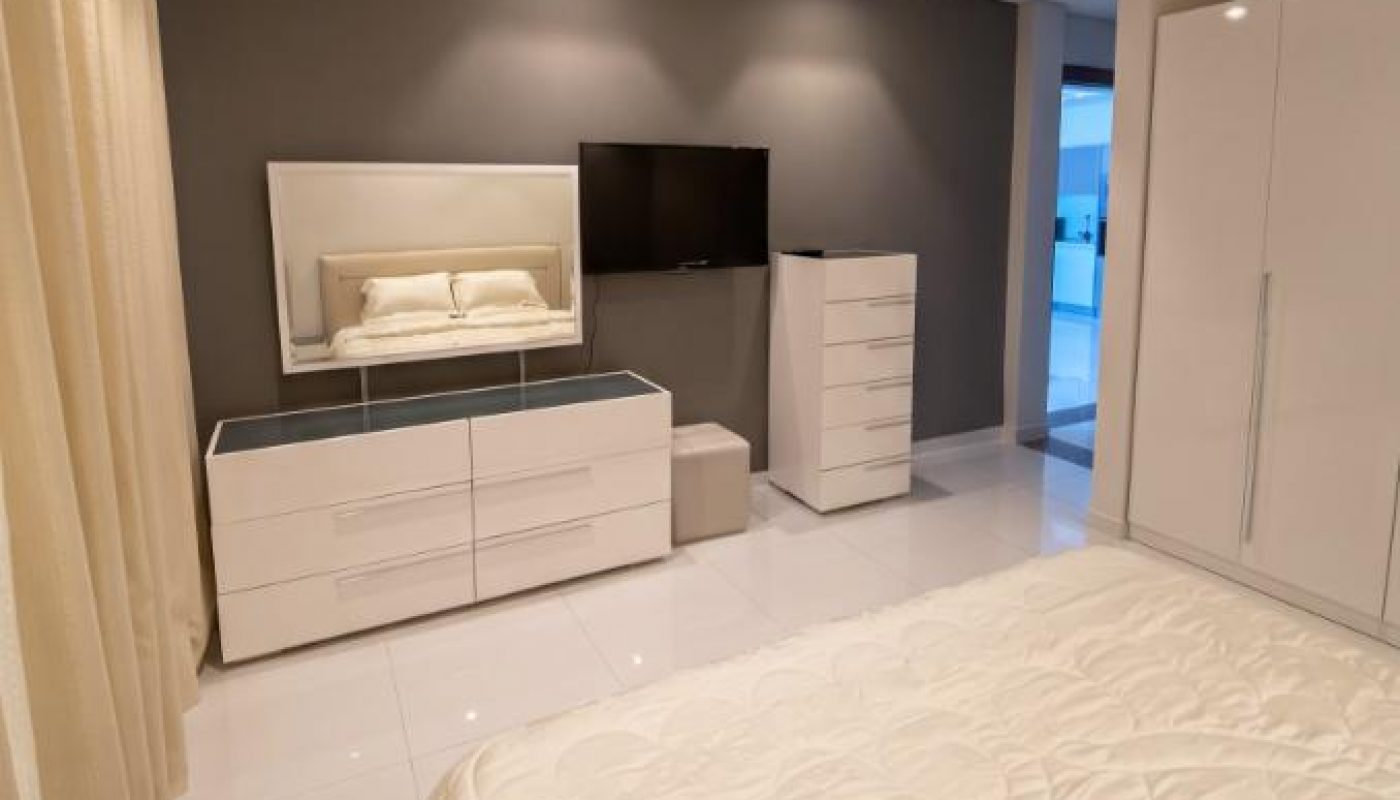Modern bedroom with a bed, white furniture, wall-mounted TV, large mirror, and neutral tones. The room features a minimalist design with bright lighting and tiled floors.