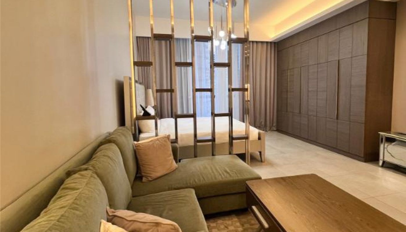 A modern studio apartment with a green sectional sofa, wooden coffee table, partition bars, bed, and built-in wardrobes. Warm lighting enhances the neutral color palette.
