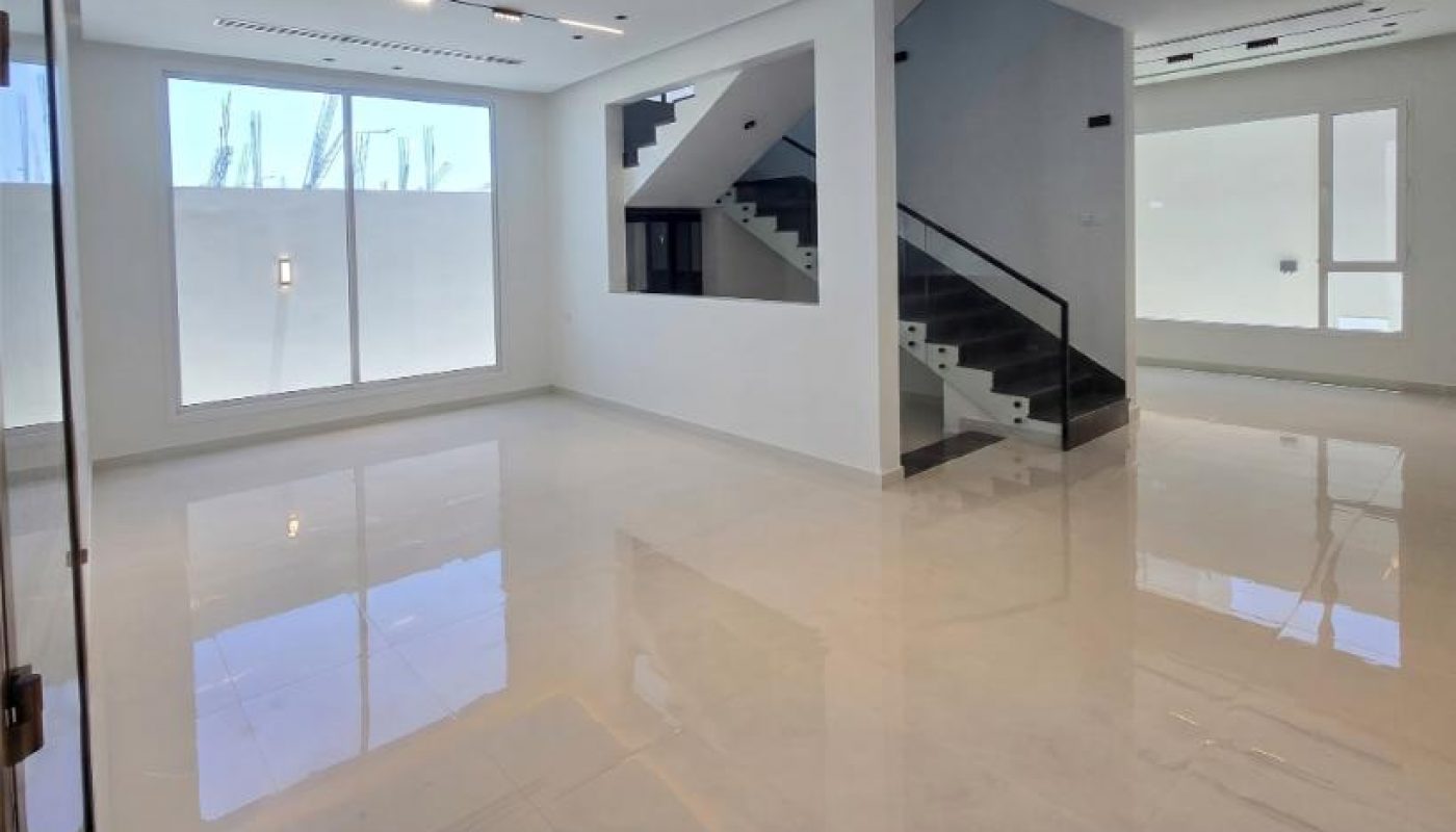 A spacious, modern, empty room with white glossy floors, large windows, and a staircase leading to an upper level.