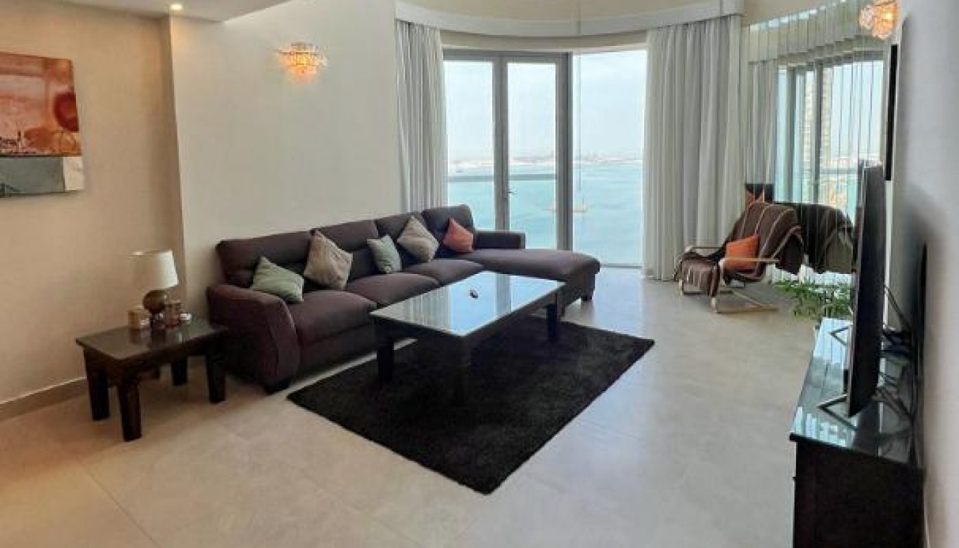 Spacious living room with a dark sofa, coffee table, TV, and large windows overlooking water, featuring beige tiled floors and modern decor.