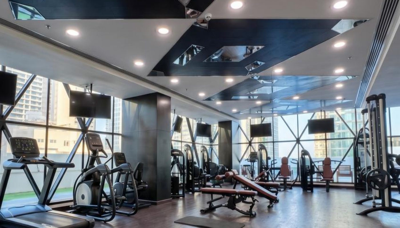 Modern gym with treadmills, elliptical machines, weight benches, and large windows; wood flooring and geometric ceiling design with recessed lighting.