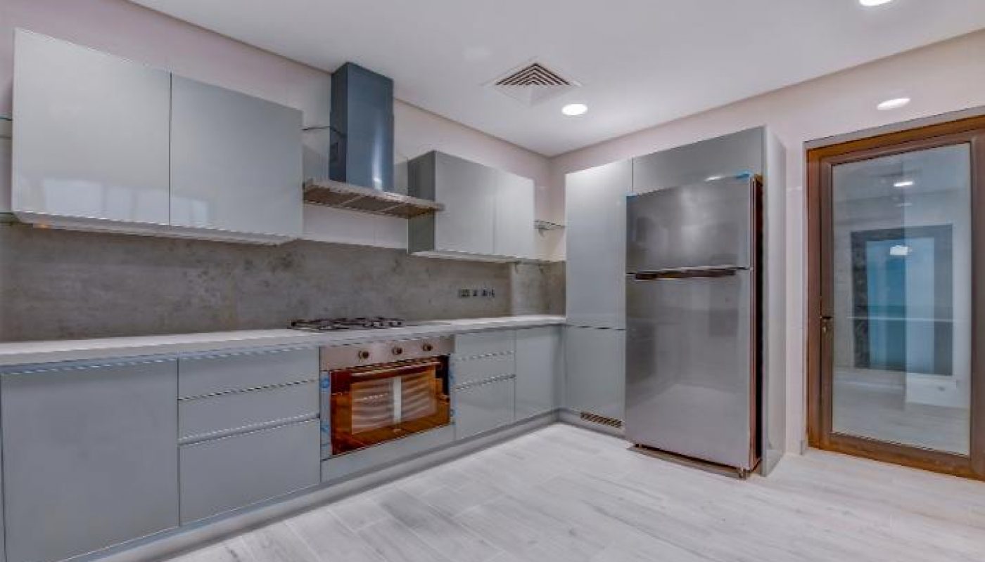 Modern kitchen with light gray cabinets, stainless steel appliances, built-in oven, and light wood flooring under recessed ceiling lights.