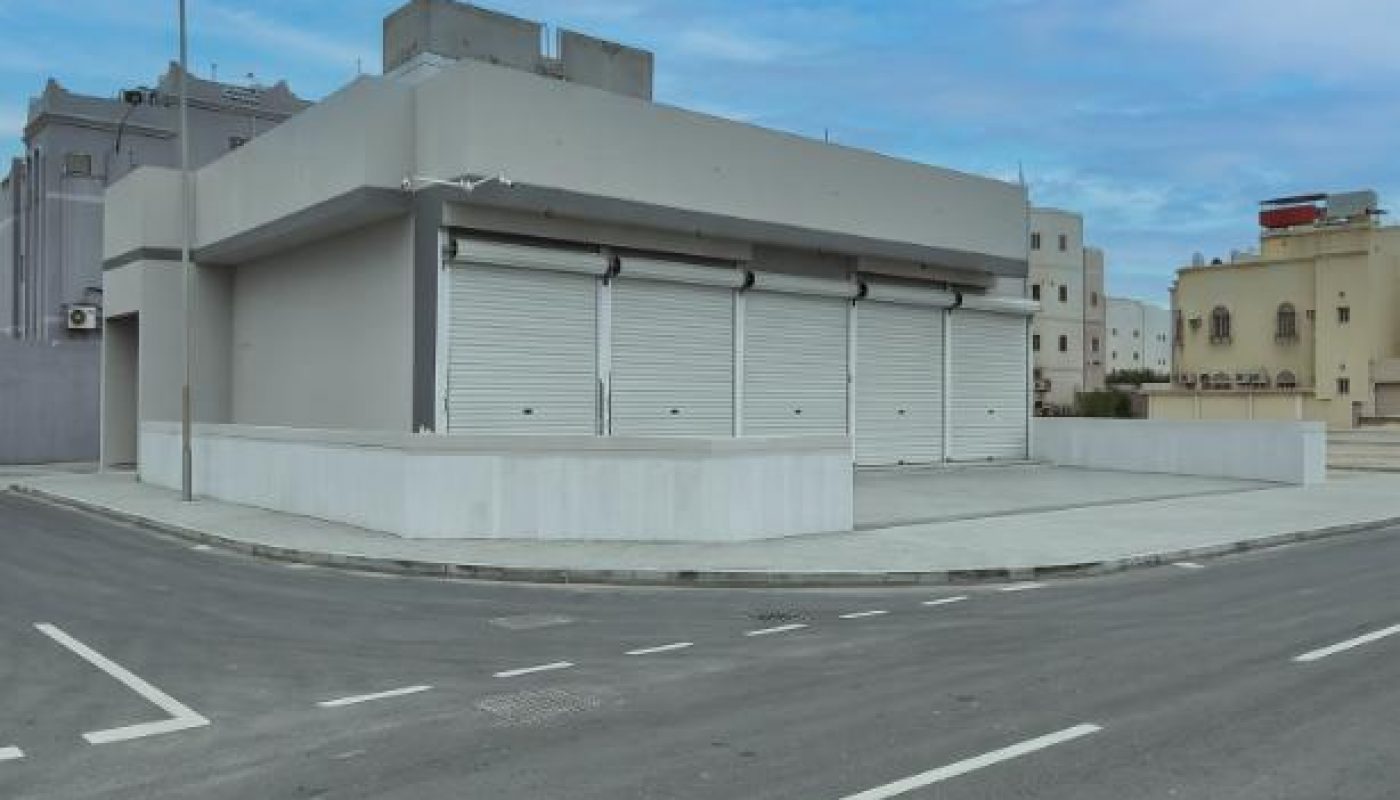 A vacant building with three closed white garage doors sits on a corner lot next to an empty street under a blue sky.